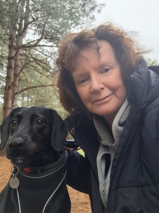 Corrinne, a previous winner, smiling next to her black labrador dog, on a woodland walk.